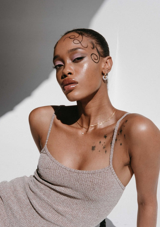 Woman with artistic face makeup and star tattoos, wearing a beige tank top, poses confidently in natural light. Fashion, beauty, and style photography.