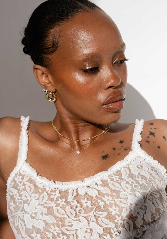 Woman in white lace top with star tattoos, wearing gold hoop earrings and layered necklaces, looking down. Fashion portrait, minimalist style.