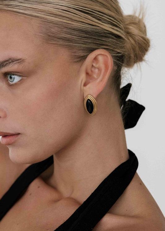 Woman wearing elegant black and gold oval earrings, hair in a bun, black halter top. Fashion accessory, jewelry close-up, minimalist style.