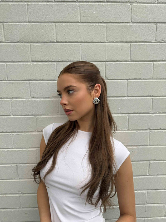 Young woman with long brown hair and white top, standing against a white brick wall, wearing large silver earrings. Fashion, hairstyle, portrait.