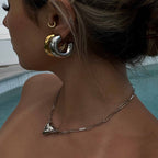 Woman wearing chunky gold and silver hoop earrings, silver chain necklace with triangle pendant, by a pool. Fashion jewelry, trendy accessories.