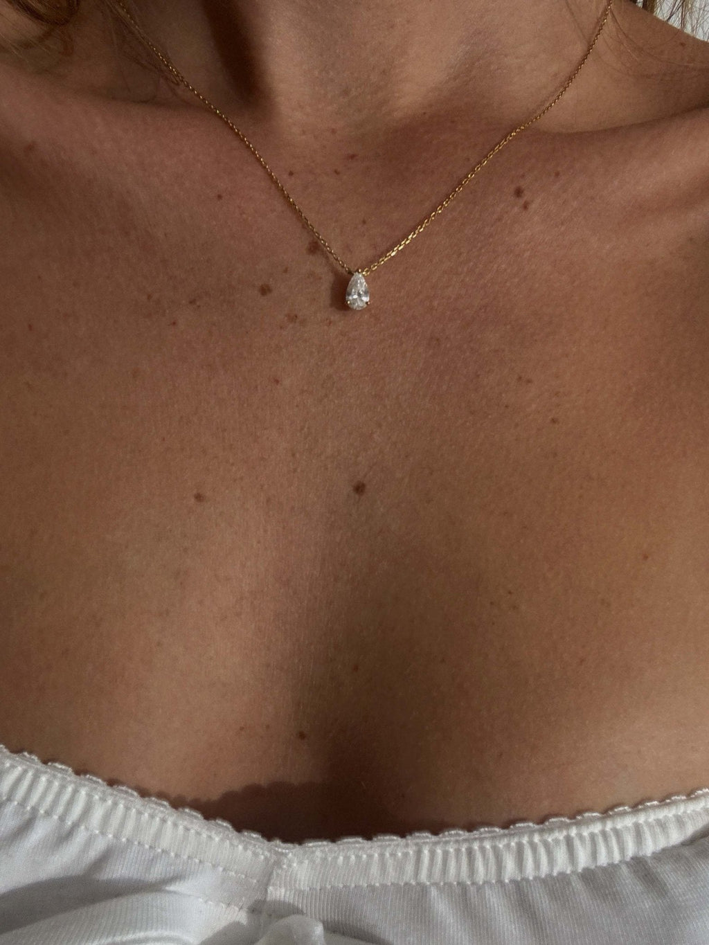 Gold necklace with a teardrop diamond pendant on a woman's neck, wearing a white off-shoulder top. Elegant jewelry, minimalist fashion accessory.