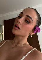 Woman with slicked-back hair and a purple orchid behind her ear, wearing gold hoop earrings and layered necklaces, posing indoors.