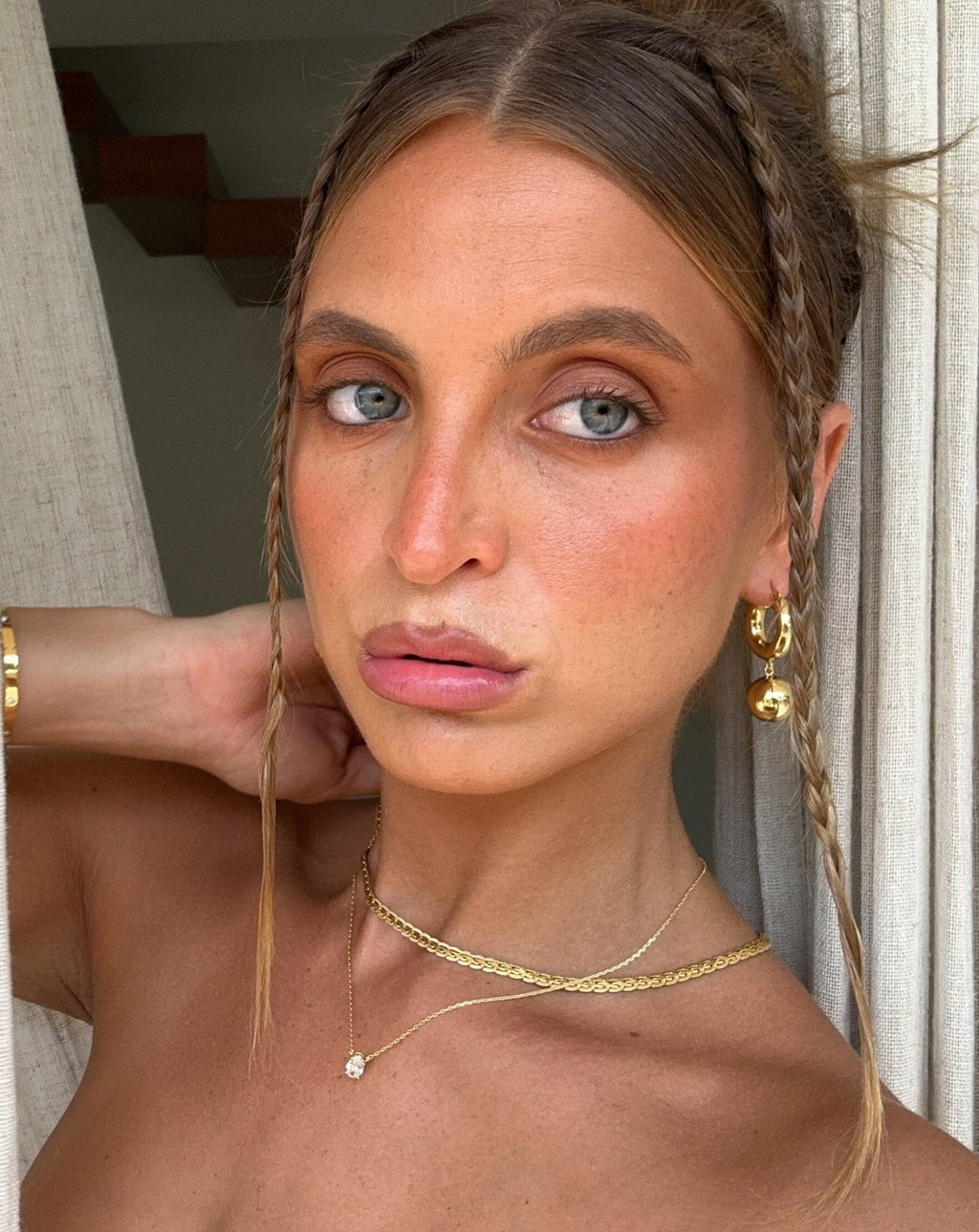 Young woman with braided hairstyle, wearing gold jewelry and natural makeup, poses indoors. Fashion, beauty, and hairstyle inspiration.