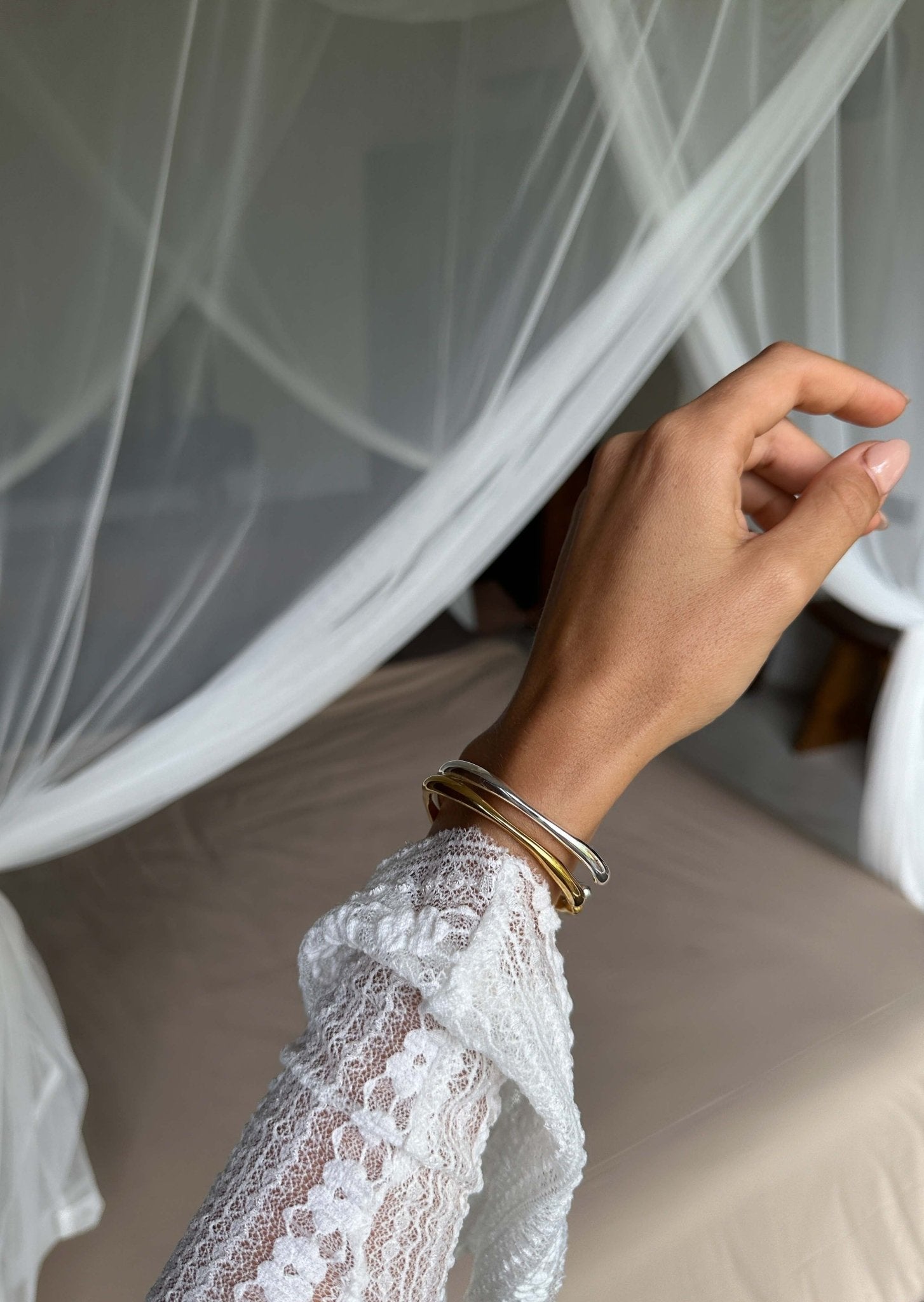 Hand with gold and silver bangles in front of a white lace canopy bed, showcasing elegant jewelry and bedroom decor.