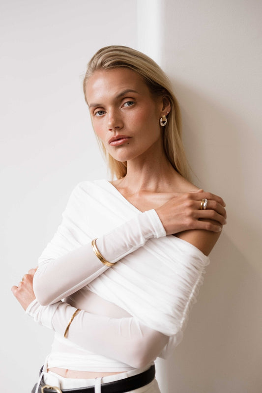 Blonde woman in white off-shoulder top, gold hoop earrings, and bracelets, leaning against a white wall, exuding elegance and modern fashion style.