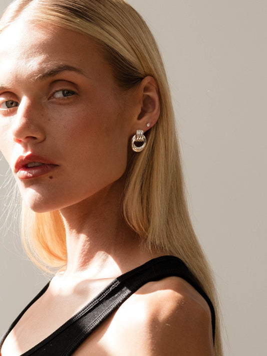Blonde woman wearing elegant gold hoop earrings, black top, and natural makeup, posing against a neutral background. Fashion jewelry portrait.