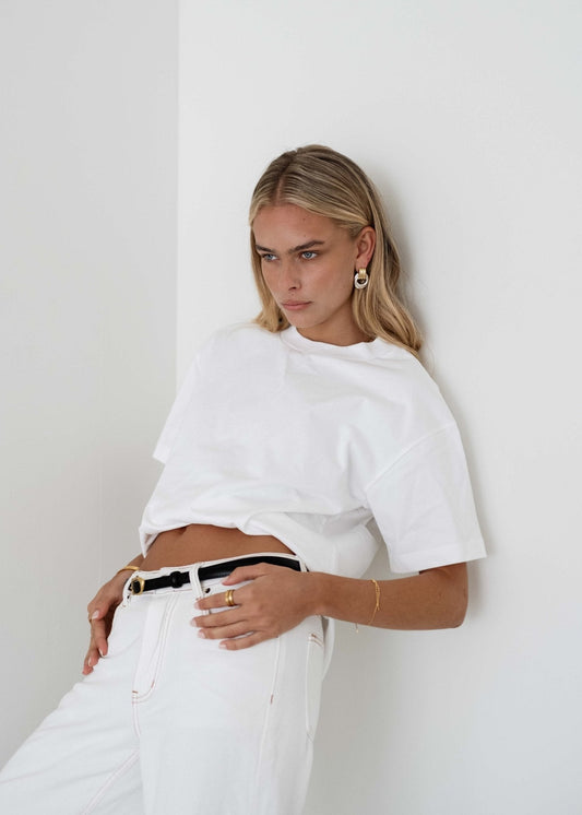Woman in white t-shirt and jeans leaning against a wall, wearing gold jewelry and a black belt. Fashionable casual outfit, minimalist style.