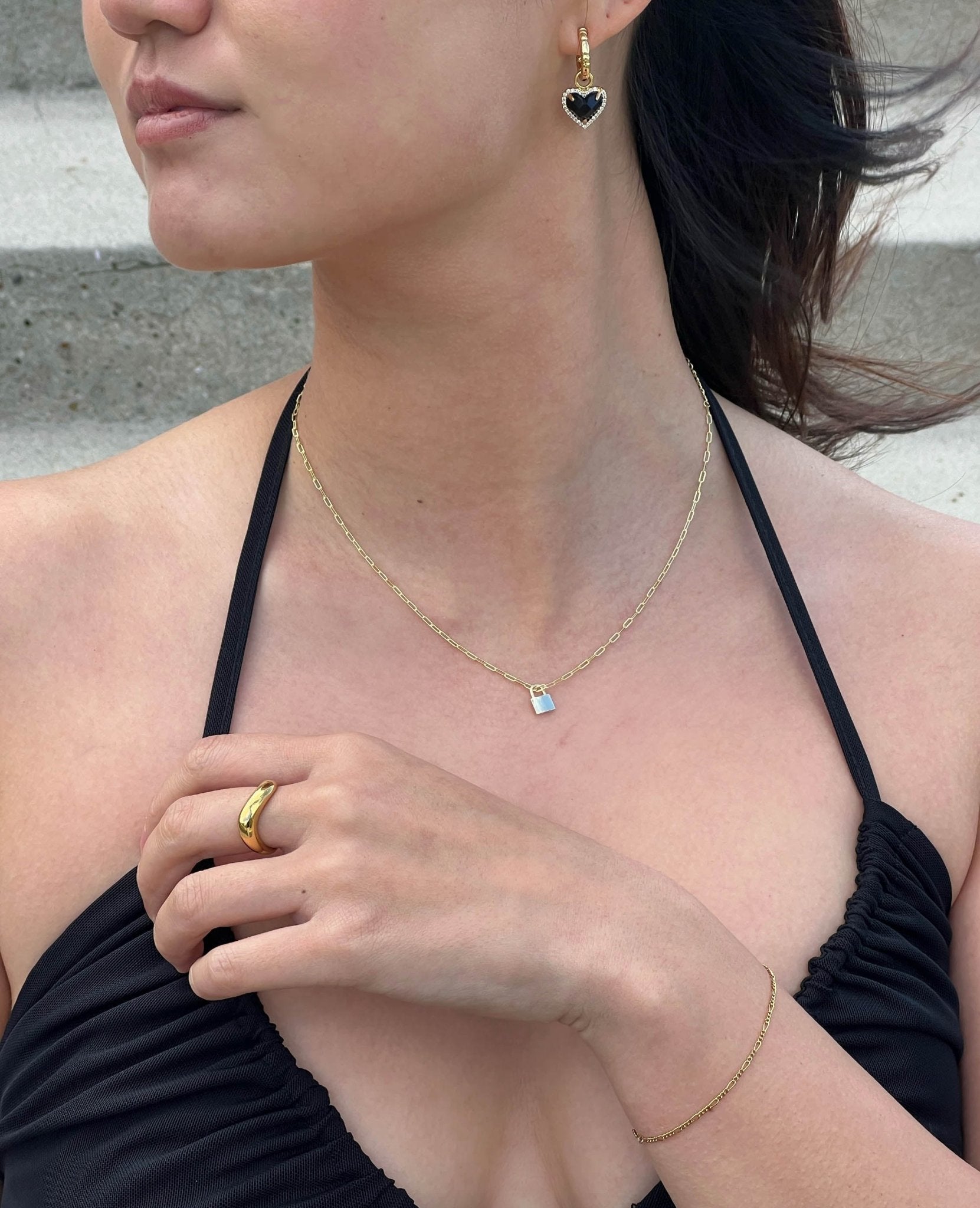Woman wearing gold jewelry: heart-shaped earrings, a pendant necklace, a ring, and a bracelet, paired with a black halter top.
