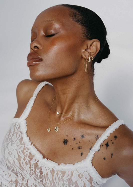 Woman in white lace top with gold jewelry and star tattoos on shoulder, eyes closed, showcasing elegant fashion and beauty.