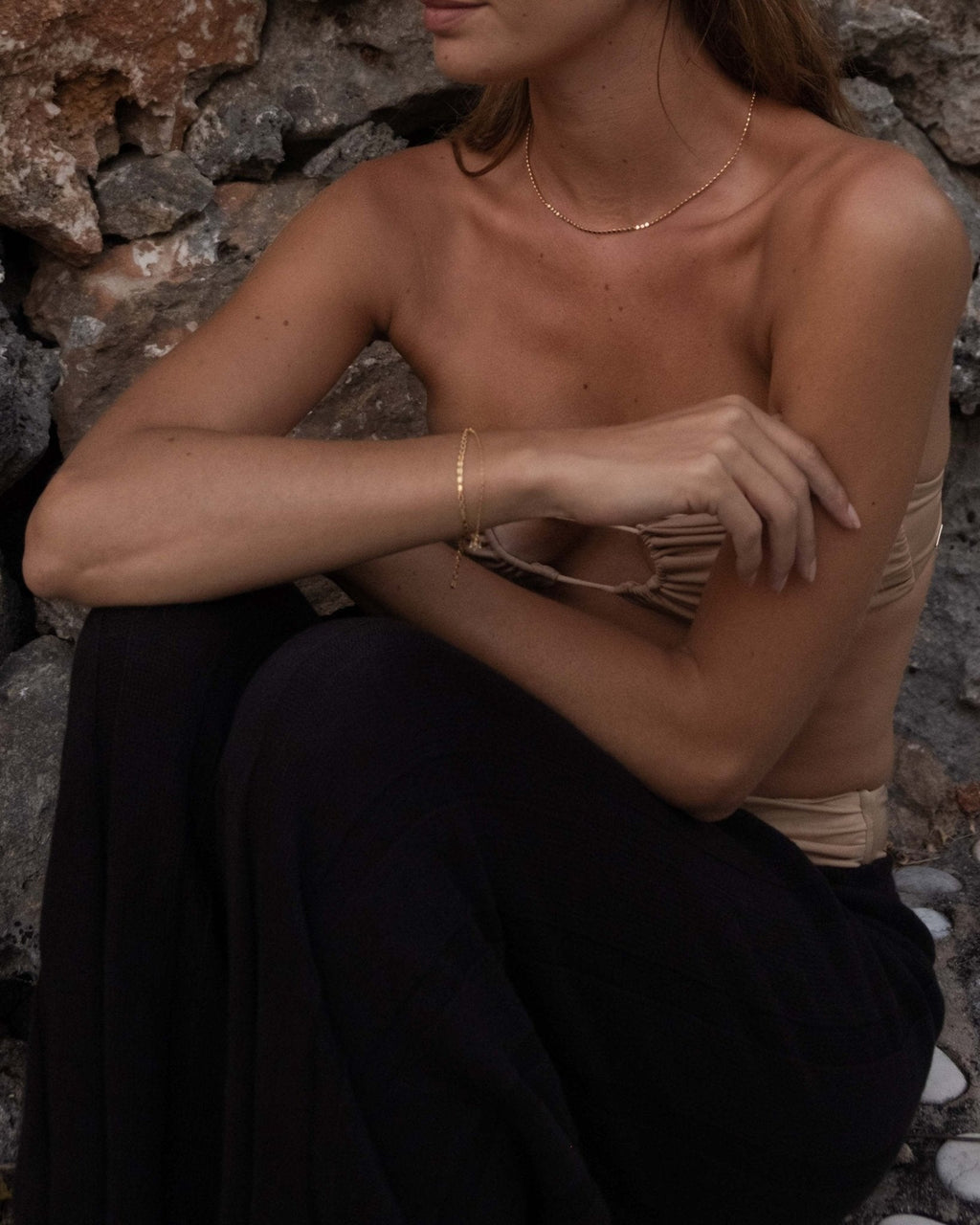 Woman in strapless top and dark pants sitting against a stone wall, wearing delicate gold jewelry; fashion, summer style, minimalist accessories.