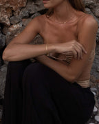 Woman in strapless top and dark pants sitting against a stone wall, wearing delicate gold jewelry; fashion, summer style, minimalist accessories.