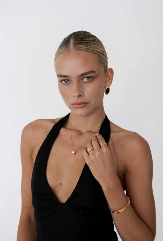 Woman in black halter dress with gold jewelry, including earrings, necklace, rings, and bracelet, against a plain white background.