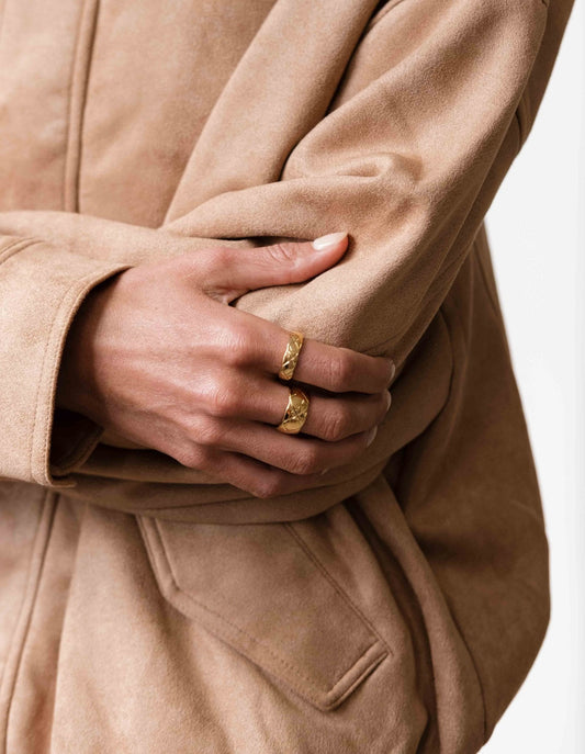 Close-up of a person wearing a beige suede jacket, showcasing two gold rings on their fingers. Fashionable jewelry and outerwear style.