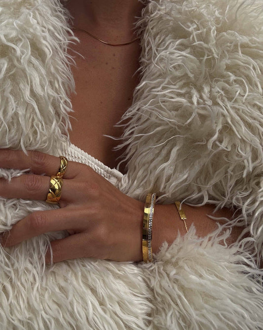 Luxurious gold jewelry on a hand wearing a fluffy white faux fur coat; features gold rings and bracelets, highlighting fashion and elegance.