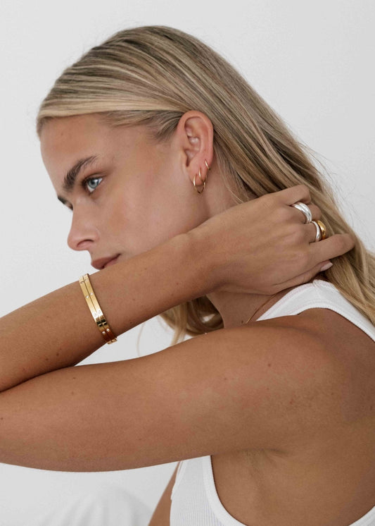 Woman wearing gold bangles, hoop earrings, and silver rings, showcasing minimalist jewelry style. Blonde hair, white tank top, side profile.