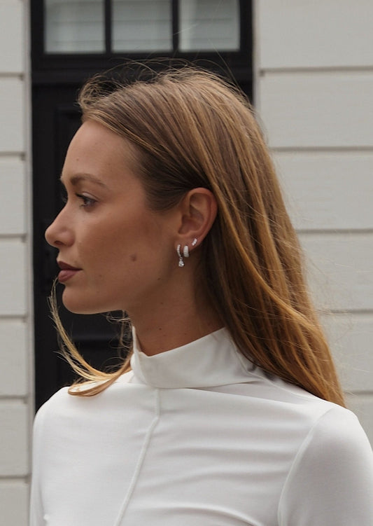 Woman in white turtleneck with long hair, wearing silver hoop earrings, standing outdoors against a white building background.