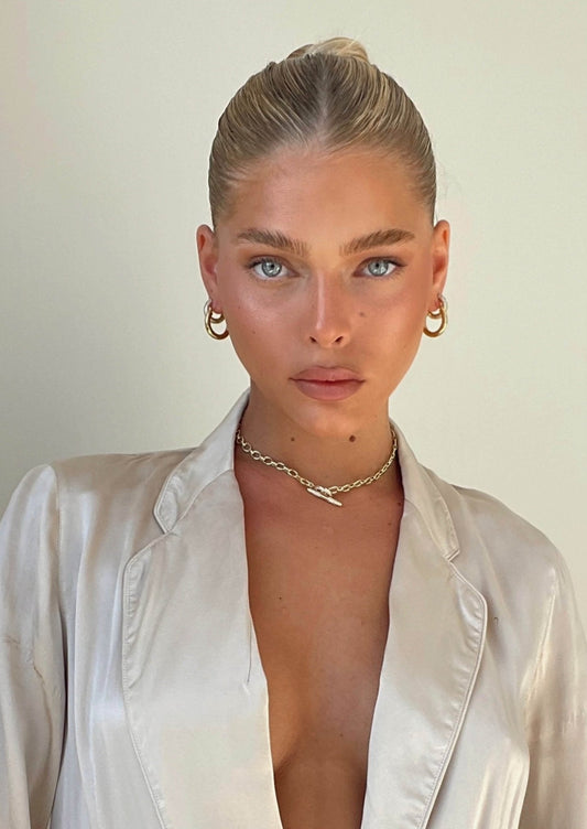 A person with blonde hair in a sleek bun, wearing a satin beige blazer, gold hoop earrings, and a chain necklace, posing against a neutral background.
