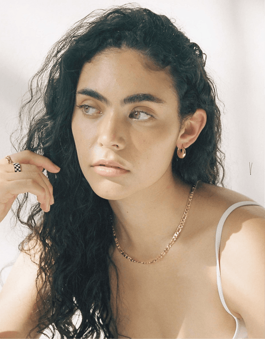Young woman with long curly hair wearing gold jewelry, including a necklace and hoop earrings, gazes thoughtfully. Natural light highlights her features.