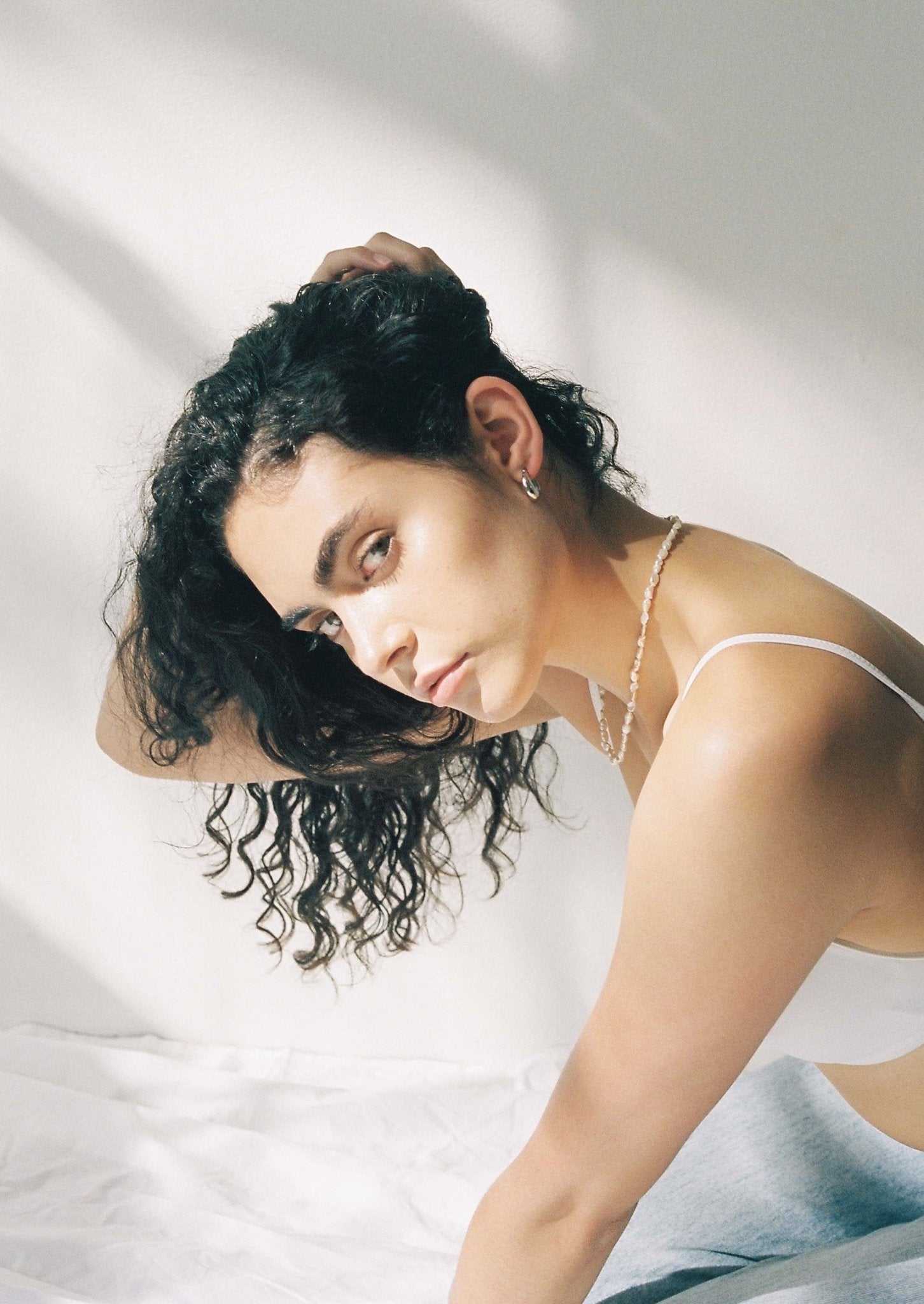 Young woman with curly hair and hoop earrings poses in natural light, wearing a white tank top and necklace, showcasing minimalist fashion style.