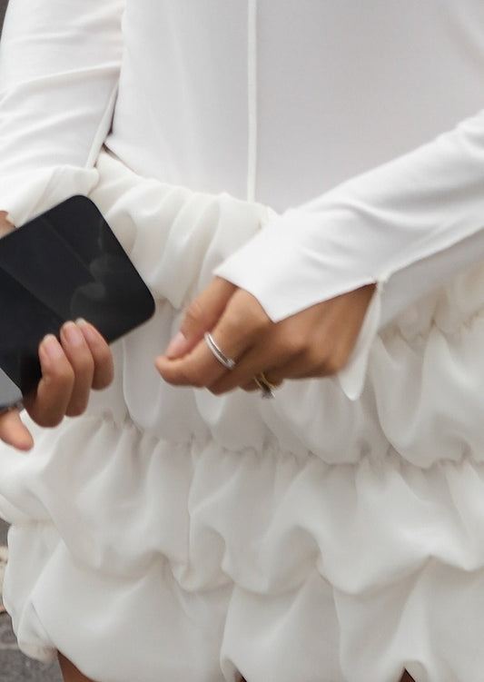 White ruffled dress with long sleeves, person holding a smartphone, elegant fashion, close-up detail, modern style, outdoor setting.