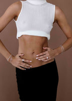 Woman wearing a white ribbed crop top and black skirt, showcasing minimalist silver jewelry on hands and wrists against a brown background.