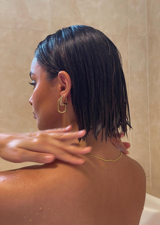 Woman with wet short hair in shower, wearing gold earrings and necklace, touching neck. Bathroom setting, focus on jewelry and hairstyle.