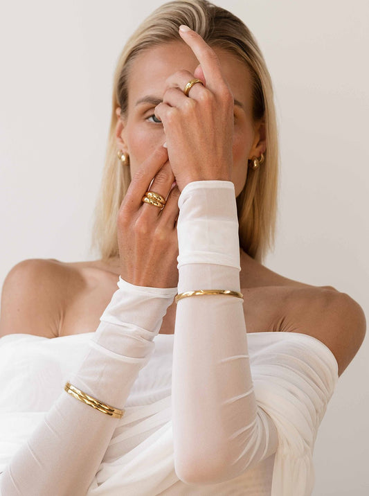 Woman wearing elegant gold jewelry, including rings and bangles, with a white off-shoulder top. Fashion accessories, minimalist style.