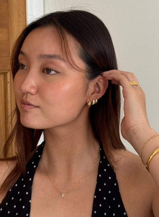 Young woman wearing gold hoop earrings, a polka dot top, and gold jewelry, including a bracelet and initial necklace, adjusting her hair.