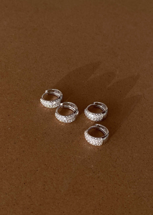Four silver hoop earrings with sparkling rhinestones on a brown surface, elegant jewelry, fashion accessories, minimalist design.