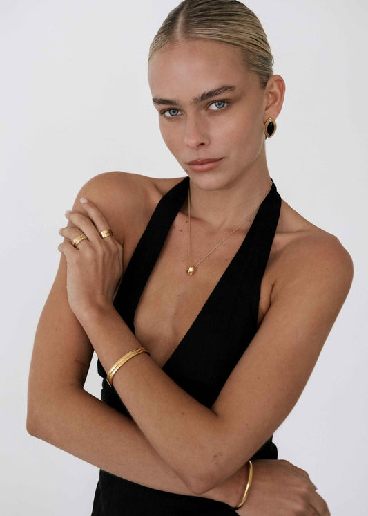 Woman in black halter dress wearing gold jewelry, including hoop earrings, necklace, rings, and bracelets, posing against a white background.