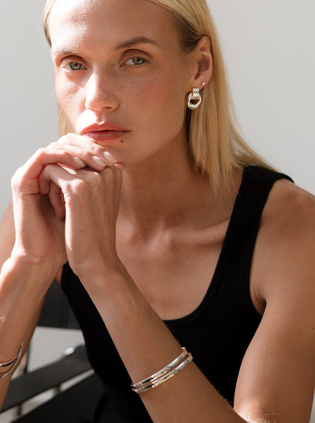 Woman wearing silver hoop earrings and bracelets, black sleeveless top, sitting thoughtfully. Fashion jewelry, minimalist style, natural light.