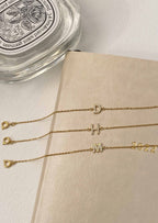 Gold initial bracelets with letters D, H, M, and numbers 2022 on a beige surface, next to a Diptyque candle lid. Elegant jewelry display.
