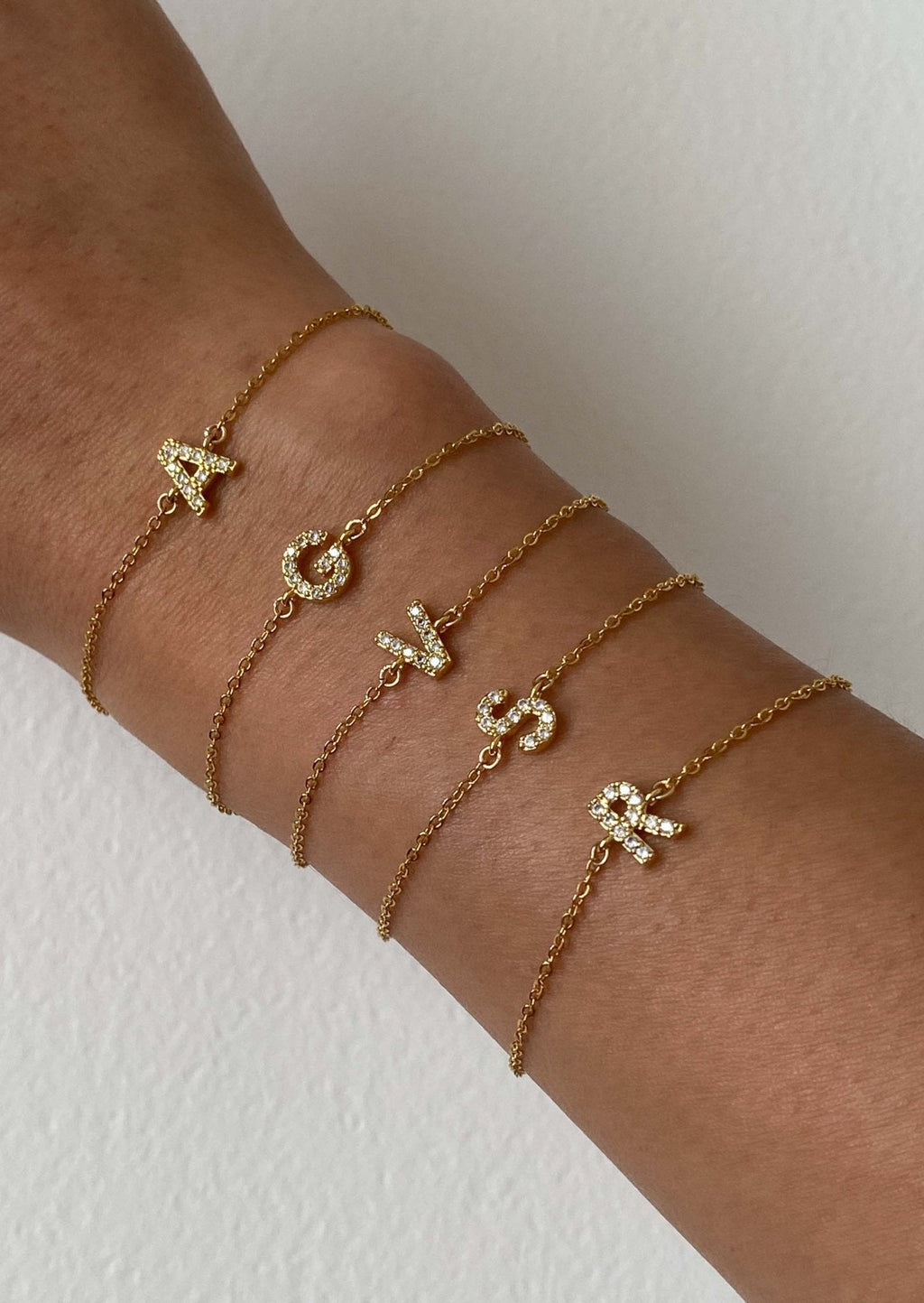 Gold initial bracelets with letters A, C, V, Y, R on a wrist, featuring delicate chains and sparkling letter charms. Perfect personalized jewelry gift.