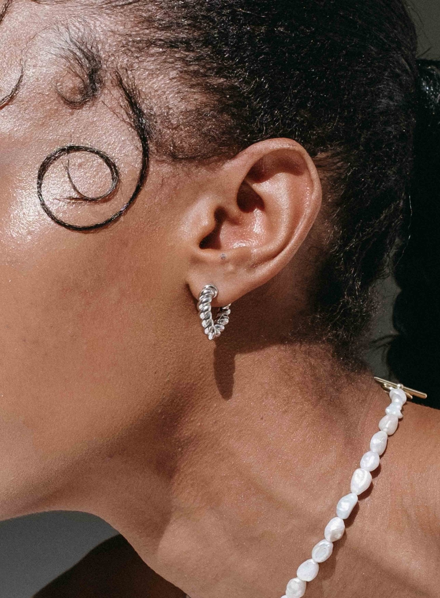 Close-up of a woman's ear wearing a twisted silver hoop earring, paired with a pearl necklace. Fashion jewelry, elegant accessories, modern style.