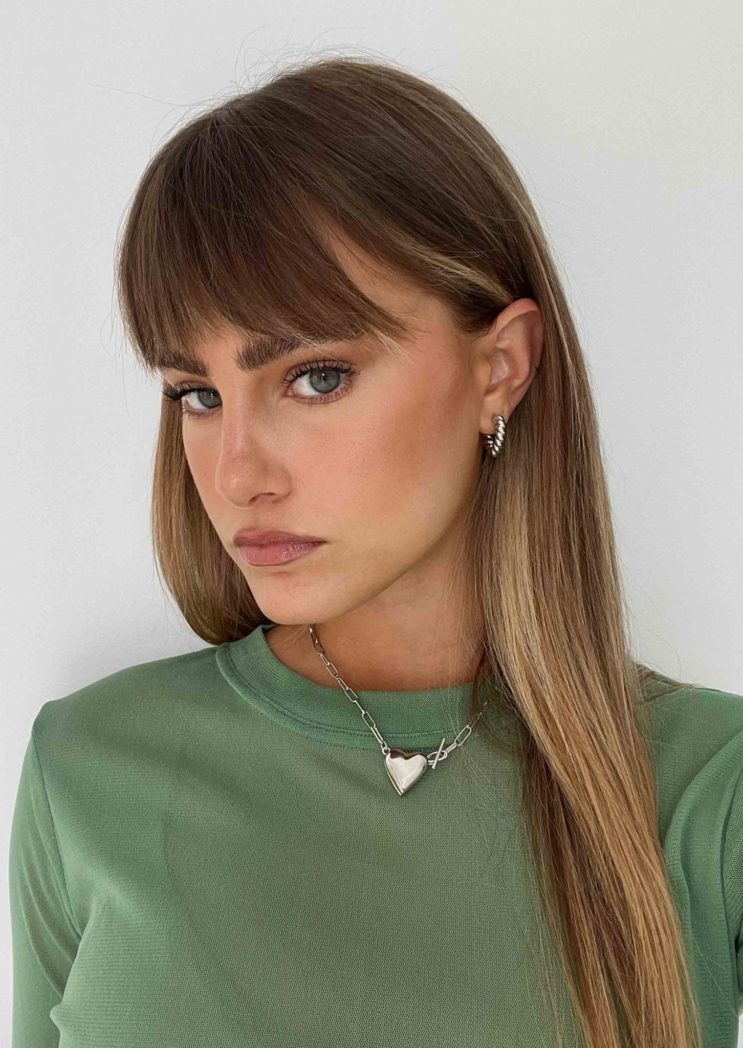 Young woman with long brown hair, wearing a green top and heart-shaped necklace, poses against a white background. Fashion, jewelry, portrait photography.