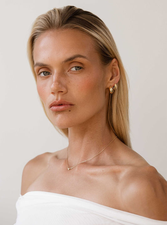 Blonde woman wearing gold hoop earrings and a delicate necklace, off-shoulder white top, neutral background. Fashion portrait, minimalist jewelry.