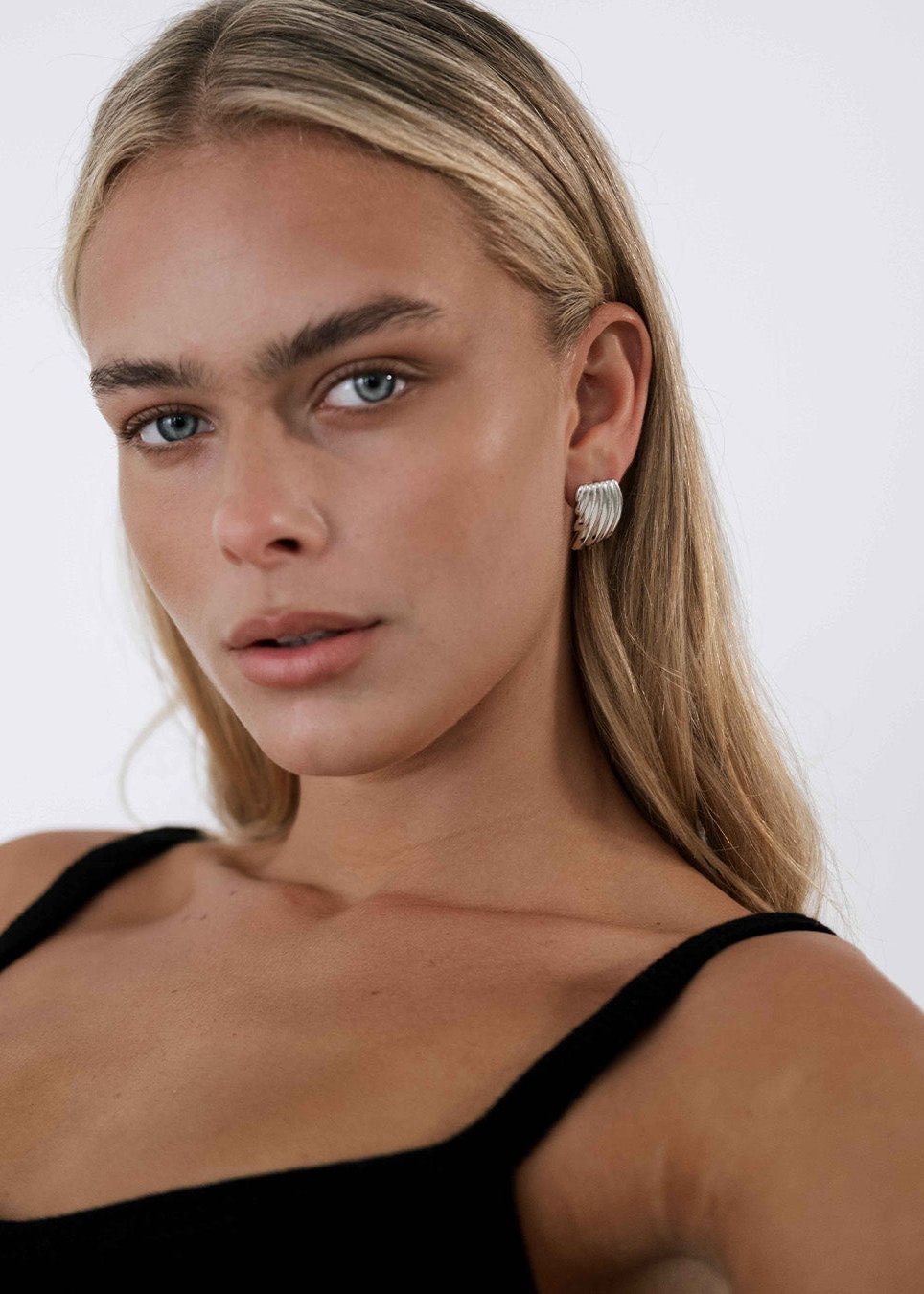 Woman wearing silver hoop earrings, black top; close-up portrait with neutral expression, blonde hair, minimal makeup. Fashion jewelry, modern style.