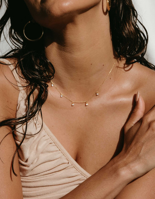 Woman wearing a gold necklace with small pendants, gold hoop earrings, and a beige ruched top, showcasing elegant summer jewelry fashion.
