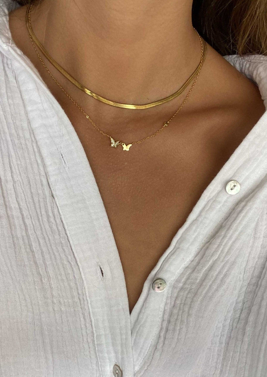 Woman wearing layered gold necklaces with butterfly pendant, white textured button-up shirt; minimalist jewelry fashion, elegant style.