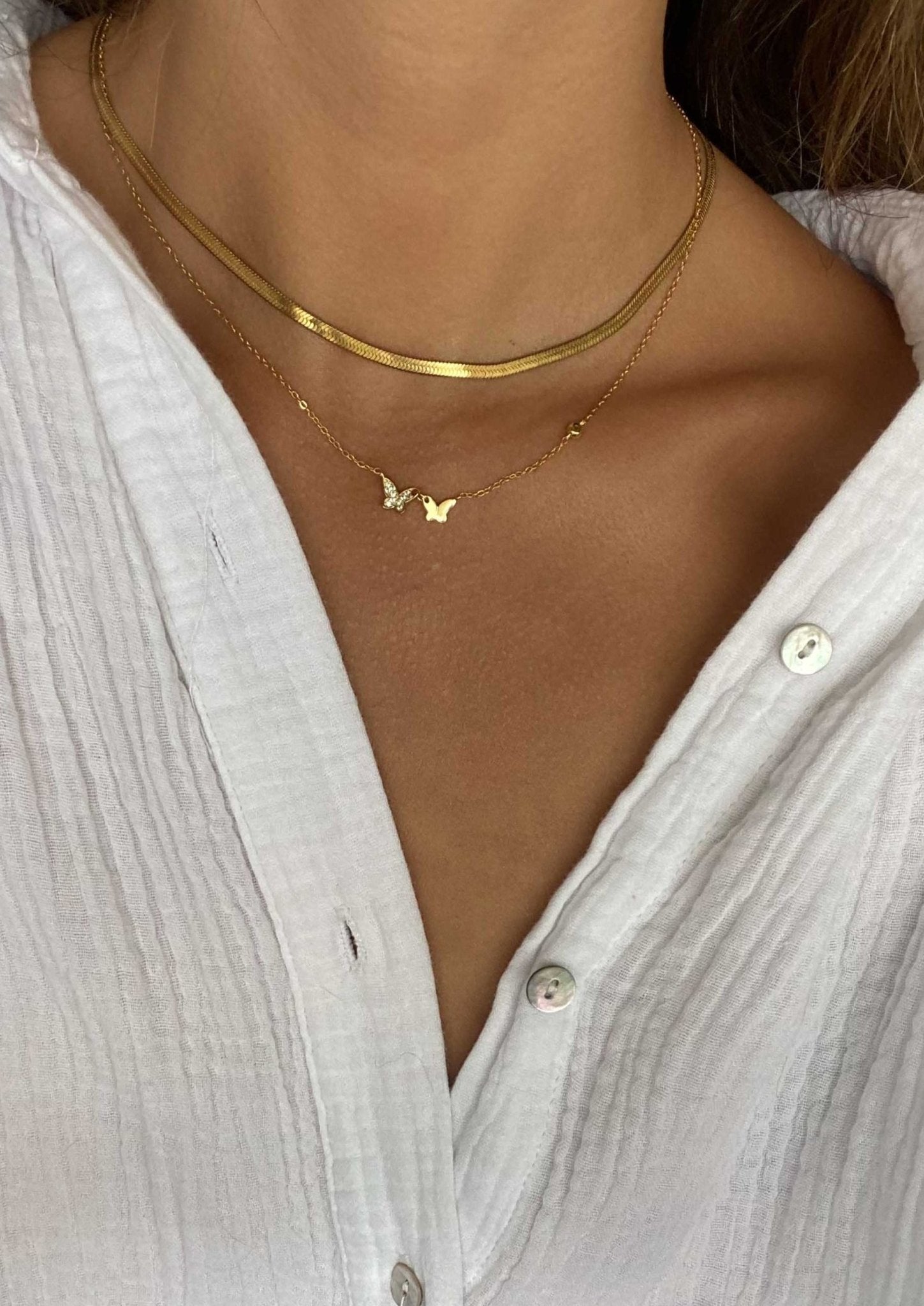 Woman wearing layered gold necklaces with butterfly pendant, white textured button-up shirt; minimalist jewelry fashion, elegant style.