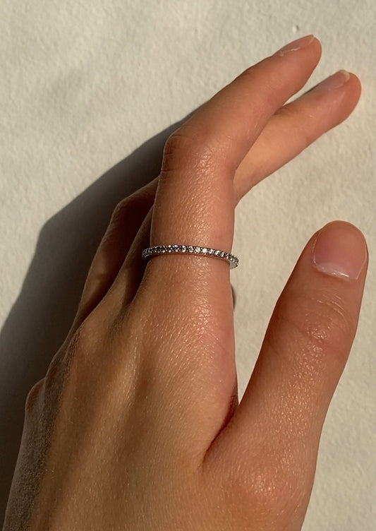 Hand wearing a delicate diamond eternity band on the ring finger, showcasing minimalist jewelry style against a soft, neutral background.